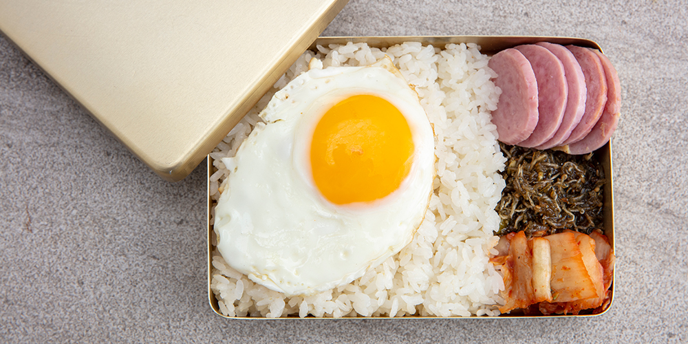 양은 도시락통에 닮긴 밥, 분홍소세지, 멸치볶음, 김치와 위에 올려진 계란 프라이